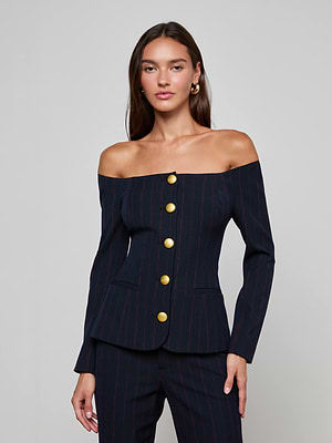 A woman exudes confidence in the L'AGENCE Alice Blazer Top, featuring an off-the-shoulder pinstripe design with bold gold buttons, posed against a simple background and staring directly at the camera with a calm expression.
