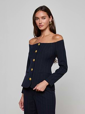 A woman poses against a plain background in the L'AGENCE Alice Blazer Top, a navy pinstripe off-the-shoulder piece with gold buttons. She wears gold hoop earrings, has long brown hair, and maintains a neutral expression.