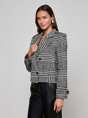 A woman with long brown hair wears the Xoey Cropped Trench Coat, black leather pants, and large round earrings, standing against a plain light gray background—a chic fall-to-winter style.