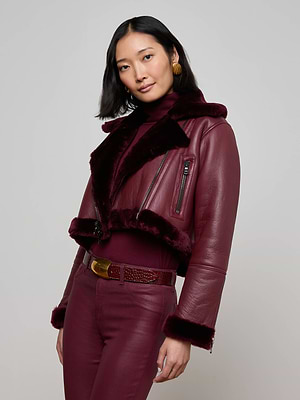 A woman with shoulder-length black hair models the Pike Shearling Leather Jacket—burgundy leather with faux fur trim—styled in a studio with a matching turtleneck, high-waisted pants, gold earrings, and a textured belt.