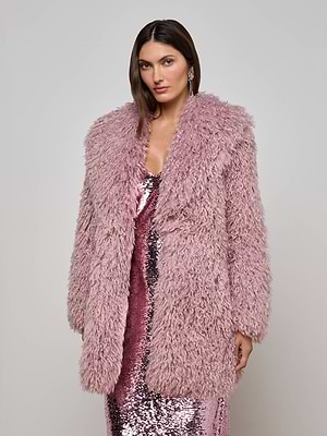 A woman wears the L'AGENCE Toira Faux Fur Coat with a shiny pink sequin dress, posing against a plain light gray background for a retro-inspired look.