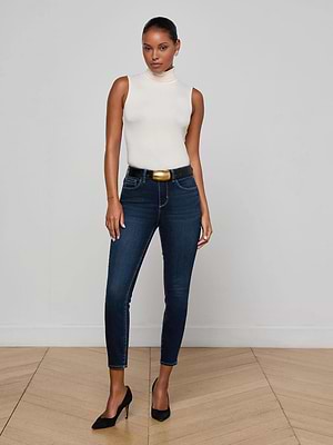 A woman stands in a minimalist room wearing L'AGENCE Margot Cropped Skinny Jeans, a sleeveless white turtleneck top, a black belt with gold buckle, and black heels on light wood flooring against a plain wall.