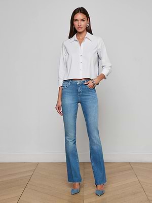A woman with long brown hair stands indoors against a light wall, wearing the L'AGENCE Ruth Straight-Leg Jean in classic stretch denim with a raw hem, paired with a white button-up shirt and blue heels, one hand in her pocket.