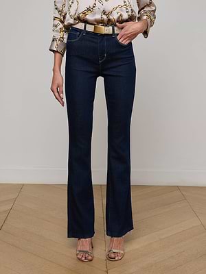 A person stands on a wooden floor wearing L'AGENCE Ruth Straight-Leg Jeans, a patterned blouse with rolled sleeves, a beige belt, and open-toe heels. The photo is cropped from the shoulders down.