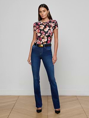 A woman with straight brown hair stands against a light wall, wearing the L'AGENCE Ruth Straight-Leg Jean in indigo, a black floral tee, black heels, and a belt. She has a neutral expression and her hands in her pockets.