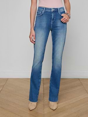 A woman models the Ruth Straight-Leg Jean, featuring a high-waisted fit and vintage wash, paired with beige pointed-toe shoes. She stands on a wooden floor against a plain white wall, with only her torso and legs visible.