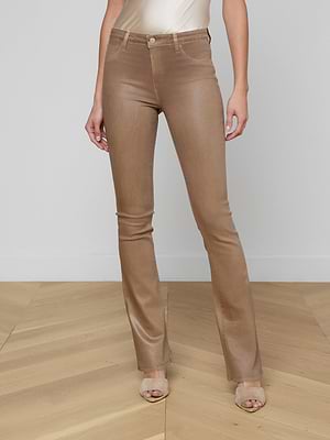 A woman stands indoors on light wooden flooring, wearing tan Selma Coated Bootcut Jeans, a light beige satin top, and open-toe heels. Her arms rest by her sides against a plain wall backdrop.