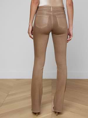 A person stands facing away from the camera, wearing Selma Coated Bootcut Jeans in light brown with beige high heels on a wooden floor, set against a plain light-colored wall.