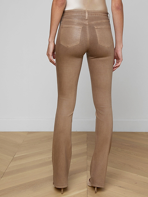 A person stands facing away from the camera, wearing Selma Coated Bootcut Jeans in light brown with beige high heels on a wooden floor, set against a plain light-colored wall.