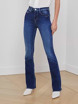 A person wears the L'AGENCE Selma Bootcut Jean in dark blue stretch denim with white heeled boots, standing against a white wall and wooden floor, hands at their sides. The top of their torso is partially visible.
