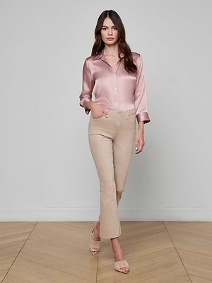 A woman with long brown hair walks on a wooden floor, wearing a light pink satin blouse, beige open-toed heels, and the neutral tan Kendra Cropped Flare Jean against a plain light-colored wall.