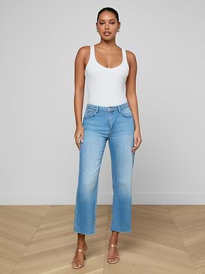 A woman stands against a plain wall wearing a white sleeveless tank and the Wanda Cropped Wide-Leg Jean in light blue, paired with tan open-toed heels. Facing forward with relaxed arms, she radiates effortless Parisian style.