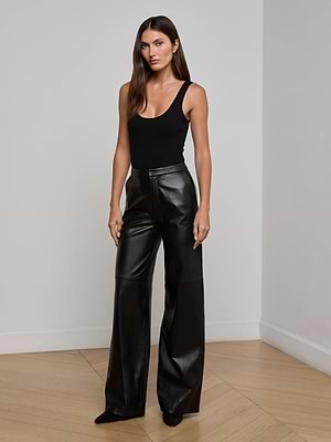 A woman with long brown hair wears the L'AGENCE Livvy Leather Trousers, a high-waisted, straight-leg style, paired with a black sleeveless top. She stands indoors on a light wood floor against a plain white wall.