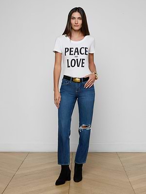 A woman stands on a wooden floor against a white wall, wearing a white PEACE AND LOVE t-shirt, Nevia Slouchy Straight-Leg Jeans with a ripped knee, a black belt, black boots, and a gold bracelet.