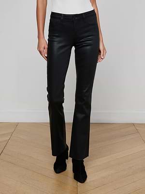 The lower half of a person is shown wearing L'AGENCE Sneeki Coated Straight-Leg Jeans and black heeled boots, standing on a wooden floor against a white wall with arms relaxed at their sides.