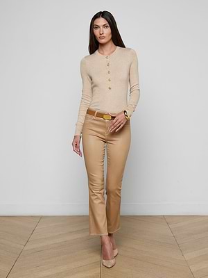 A woman with long dark hair stands against a plain white wall, wearing a beige long-sleeve top, beige heels, and the Tati Coated Bootcut Jean in tan. She poses with one hand slightly raised and a neutral expression.