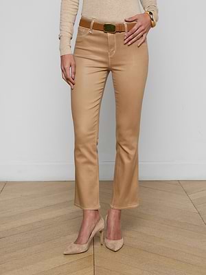 A person wearing the Tati Coated Bootcut Jean in beige with a matching belt, beige long-sleeve top, and beige heels stands on a wooden floor against a plain white wall.