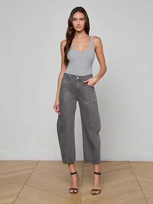 A woman with long wavy hair and a neutral expression stands indoors against a light gray wall and wooden floor, wearing a light gray tank top, Cody Cropped Barrel-Leg Jean in all-cotton denim, and black strappy heels.