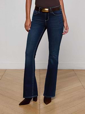 A person wearing L'AGENCE Cara Low-Rise Bootcut Jeans in dark blue with a brown belt and pointed-toe shoes stands on a light wooden floor against a plain white wall, shown from shoulders down.