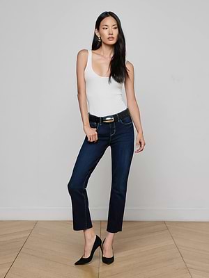 A woman with long dark hair poses in a minimal room wearing a white sleeveless top, black belt, high heels, and Aiden Low-Rise Slim-Leg Jeans. One hand rests on her hip while the other is relaxed.