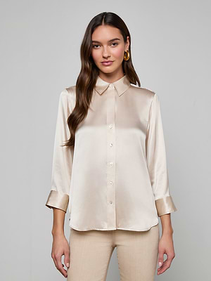 A woman with long brown hair wears the Dani Silk Blouse, a champagne-colored, shiny satin button-down with a collar and three-quarter sleeves, paired with light beige pants against a plain gray background.