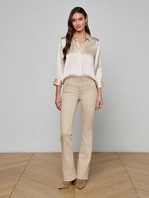 A woman stands on a wooden floor indoors, wearing the Dani Silk Blouse in champagne, beige pants, and taupe heeled boots, set against a plain light gray wall.