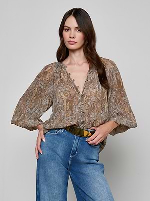 A woman stands against a plain light gray background, wearing the Teagan Blouse—a loose brown paisley print top with balloon sleeves in crinkle poly chiffon—paired with blue high-waisted jeans and her hand on her hip.