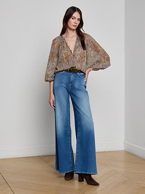 A woman poses indoors with one hand in her pocket, wearing the Teagan Blouse—a semi-sheer paisley print top with billowy sleeves—paired with wide-leg blue jeans, a brown belt, and brown pointed-toe boots against a plain white wall.