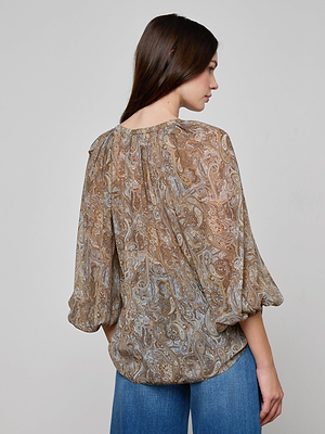 A woman with long brown hair, seen from the back, wears the Teagan Blouse—a semi-sheer paisley print crinkle poly chiffon top with three-quarter sleeves—paired with blue jeans against a plain light gray background.