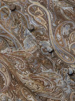 Close-up of the Teagan Blouse in brown paisley print with intricate blue and tan details, crafted from crinkle poly chiffon and featuring round fabric-covered buttons on its semi-sheer surface.