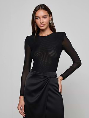 A woman with long brown hair wears the L'AGENCE Trista Bodysuit—a black mesh style with structured shoulder pads—paired with a high-waisted black satin skirt. She stands against a light gray background, looking at the camera.