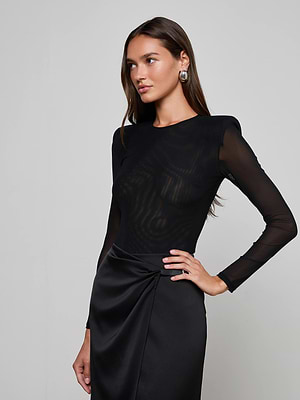 A woman models the L'AGENCE Trista Bodysuit, a sheer black mesh long-sleeve style, paired with a satin black skirt. She stands against a plain light background, posing with one hand on her hip, facing slightly to the side.