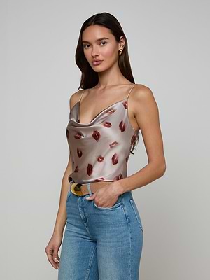 A woman with long brown hair models the Calista Satin Camisole, featuring adjustable straps and a lip print design, paired with light blue jeans. She poses with one hand on her hip against a plain light background.
