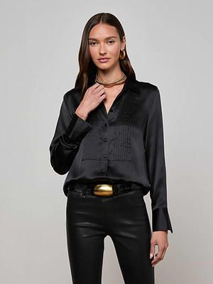 A woman with long brown hair wears the L'AGENCE Divya Silk Shirt, paired with black leather pants, gold hoop earrings, layered necklaces, and a gold belt buckle, standing against a plain light gray background.