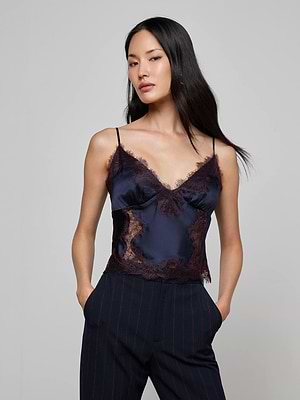 A woman with long black hair wears the L’AGENCE Kimber Silk Lace Camisole, featuring a deep V-neckline, paired with dark pinstripe pants. She stands with her hands in her pockets against a plain gray background.