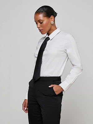 A woman with short, slicked-back hair stands sideways against a light gray background, wearing the white Blake Bodysuit and black pants with a removable tie, one hand in her pocket.
