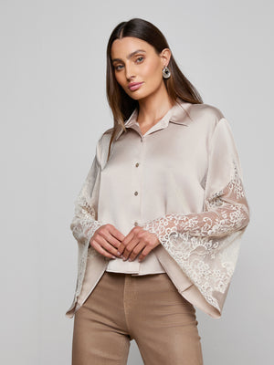 A woman with long brown hair and hoop earrings models the Capri Lace Sleeve Blouse, a silky light beige bohemian top with lace bell sleeves, paired with tan pants, against a plain light background.