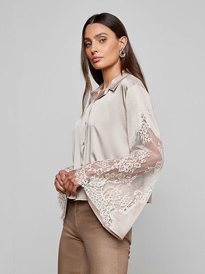 A woman with long brown hair models the Capri Lace Sleeve Blouse, a light beige bohemian top with lace bell sleeves, paired with tan pants. She stands sideways and looks at the camera against a plain gray background.