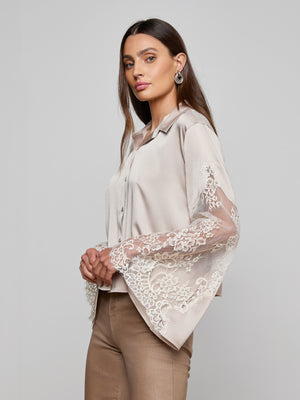 A woman with long brown hair models the Capri Lace Sleeve Blouse, a light beige bohemian top with lace bell sleeves, paired with tan pants. She stands sideways and looks at the camera against a plain gray background.