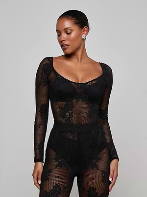 A woman wears the L'AGENCE Gwendolyn Mesh Knit Bodysuit with black undergarments, standing against a plain background. Her hair is pulled back, she looks to her left, and accessorizes with stud earrings.