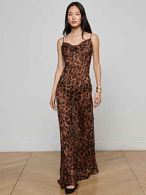 A woman poses indoors in the Christine Silk Dress by L'AGENCE, featuring a leopard print and thin straps. She pairs it with gold bracelets and black heels against a light wall and wooden floor—an ideal statement evening look.