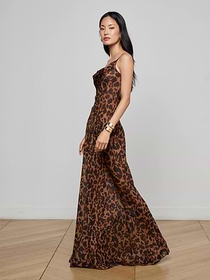 A woman in the Christine Silk Dress by L'AGENCE, a long sleeveless leopard print gown, stands sideways on a wooden floor against a white wall. She has straight dark hair and gold bracelets—an effortlessly chic evening look.