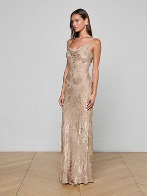 A woman poses against a plain light-colored wall on a wooden floor, wearing the Christine Burnout Satin Gown—a gold metallic dress with thin straps, a draped neckline, and a floral pattern.