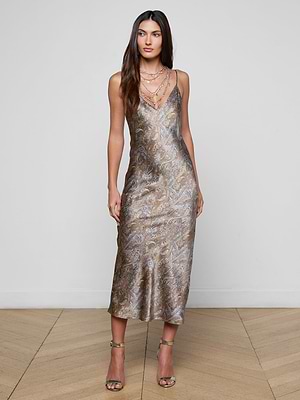A woman wears the Seridie Silk Slip Dress, featuring a metallic paisley print, spaghetti straps, and a v-neckline, paired with layered necklaces and strappy high-heeled sandals on a wooden floor against a plain background.
