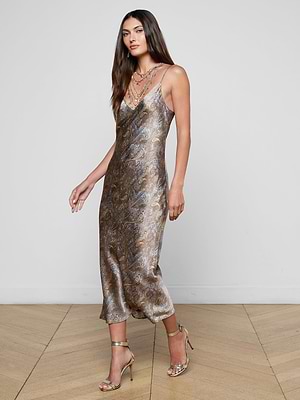 A woman with long brown hair wears the Seridie Silk Slip Dress—a sleeveless metallic paisley print slip—styled with gold high-heeled sandals and layered necklaces, standing on a wooden floor against a plain white wall.