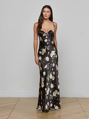 A woman with long brown hair poses against a plain light gray wall, wearing the L'AGENCE Sayla Silk Gown—a long black dress with thin straps and a yellow floral print—on a wooden floor.