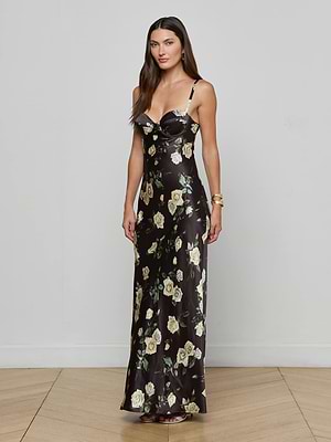 A woman with long brown hair wears the Sayla Silk Gown by L'AGENCE—a black, floor-length satin dress with light yellow rose prints, thin straps, and a sweetheart neckline. She stands indoors on a light wood floor against a gray wall.