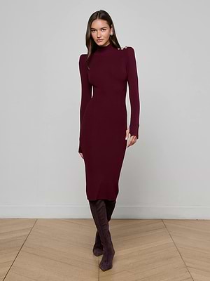 A woman stands against a light gray wall, wearing the L'AGENCE Larelle Midi Dress—a maroon ribbed knit with long sleeves, a mock neck, and shoulder buttons—paired with matching knee-high boots on a wooden floor.