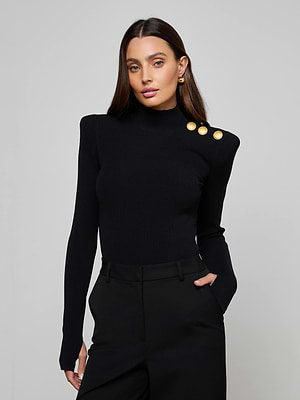 A woman with long brown hair wears the L'AGENCE Jennis Mock-Neck Bodysuit, featuring three gold shoulder buttons, paired with black pants. She stands against a plain light gray background, one hand in her pocket.