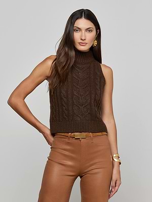 A woman with long brown hair wears the L'AGENCE Bellini Cable-Knit Turtleneck, tan leather pants, gold earrings, and a gold bracelet, standing against a plain light gray background.